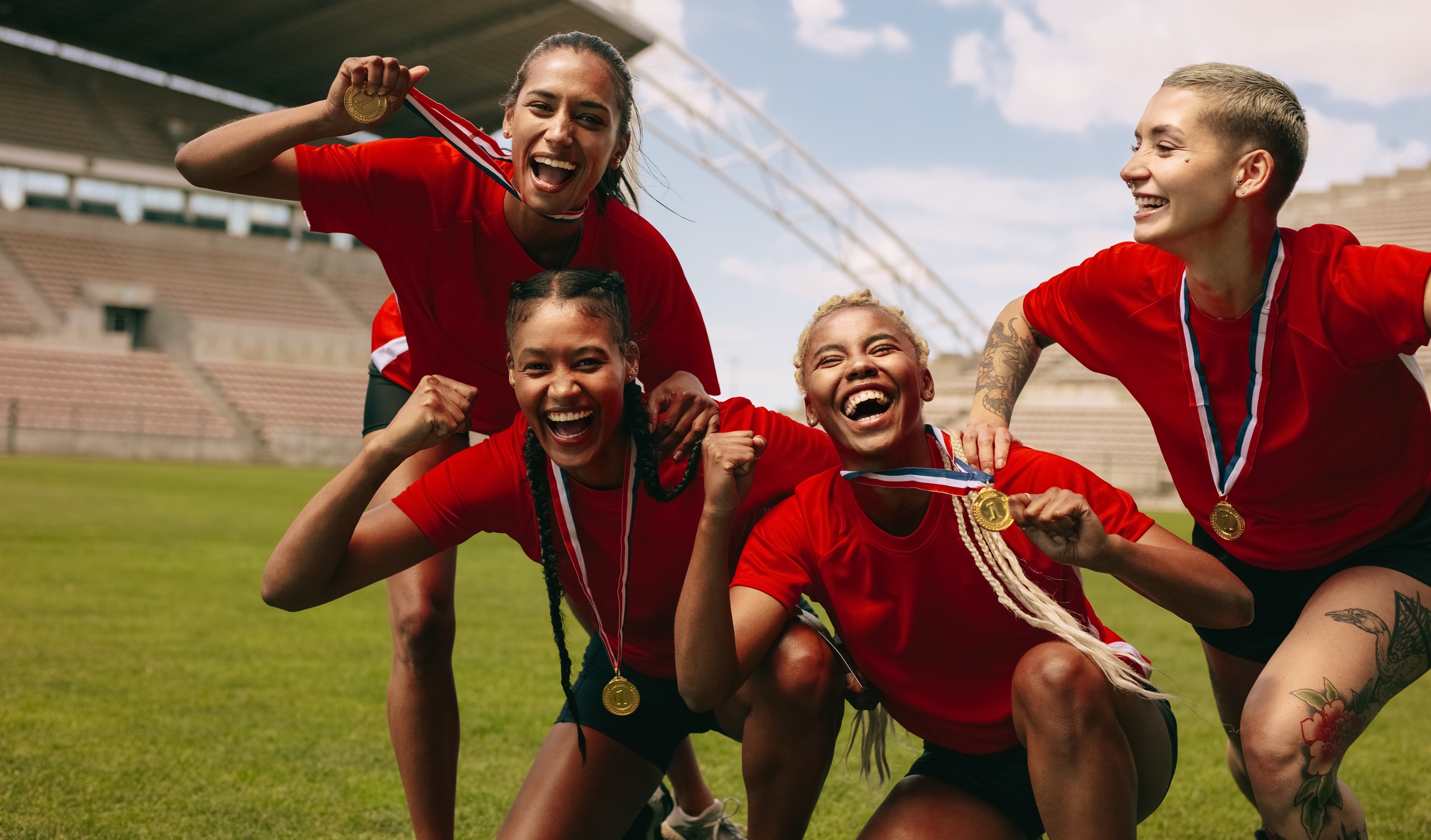 Four Smiling Female Athletes in Red Shirts and Black Shorts Standing on a Soccer Field with Medals Around their Necks
